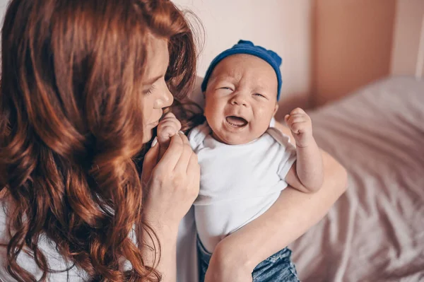 Close up portrait of happy young mom holding her crying baby in blue jeans and white t-shirt and cap. Young happy family, mother playing with cute emotional little newborn child son in the bedroom.