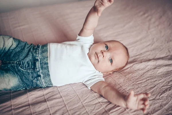 可爱的小男孩躺在卧室粉红色的毛毯上 新生儿 婴儿在床上保持清醒 穿着蓝色牛仔裤 蓝色帽子和白色T恤的有感情的新生儿的肖像 — 图库照片