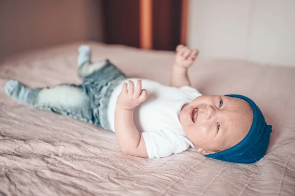 可爱的小男孩在卧室里躺在粉红色的毯子上哭泣 新生儿 婴儿在床上保持清醒 穿着蓝色牛仔裤 蓝色帽子和白色T恤的有感情的新生儿的肖像 — 图库照片