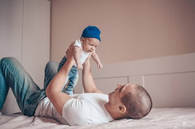 Bebeğini kot pantolon, beyaz tişört ve şapkayla tutan mutlu baba portresi. Genç, mutlu bir aile, baba yatak odasında küçük, tatlı, duygusal bir çocukla oynuyor..