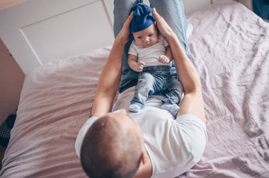 Bebeğini kot pantolon, beyaz tişört ve şapkayla tutan mutlu baba portresi. Genç, mutlu bir aile, baba yatak odasında küçük, tatlı, duygusal bir çocukla oynuyor..