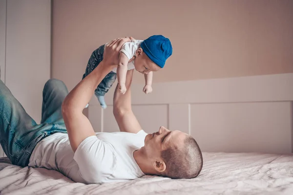 Close up portrait of happy young dad father holding his baby in blue jeans and white t-shirt and cap. Young happy family, dad playing with cute emotional little newborn child son in the bedroom.