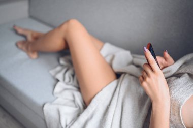 Akıllı telefon kullanan bir kadın. Gri bir kanepede, evde rahat bir battaniyeyle mükemmel ciltli ince bacaklı bir kadın. Epilasyon, sağlık, pedikür, vücut bakımı, sağlıklı tırnaklar, lazer tüy temizleme konsepti.