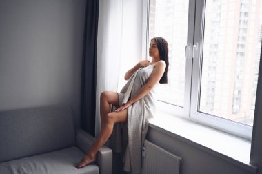 Güzel, sıska esmer kadın pencerenin kenarında oturuyor sıcak gri battaniyenin altında. Corona virüsü salgını sırasında kendini karantinaya al. COVID19 evde kalıp hayat kurtarma kavramı