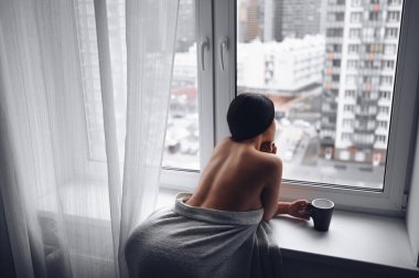 Güzel, sıska esmer kadın pencerenin kenarında oturuyor sıcak gri battaniyenin altında. Corona virüsü salgını sırasında kendini karantinaya al. COVID19 evde kalıp hayat kurtarma kavramı