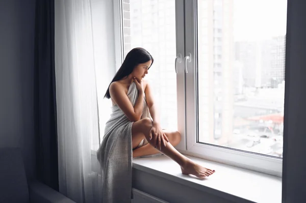 美しい退屈スレンダーなブルネットの女性は家で暖かい灰色の毛布の下で窓の横に座っている コロナウイルスパンデミックの間の自己隔離隔離 Covid19 ホームセーブライフコンセプト — ストック写真