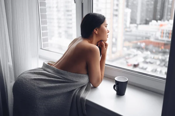 Beautiful bored slender brunette woman sitting next to window windowsill under warm grey blanket at home. Self isolation quarantine during Corona virus pandemic. COVID19 stay home save lives concept