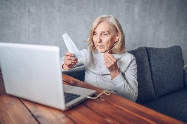 Teknoloji, yaşlılık ve insan konsepti - Coronavirus COVID19 salgını sırasında yüz maskesi takan yaşlı bir kadın evde dizüstü bilgisayarla video görüşmesi yapıyor. Evde kal konsepti