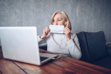 Teknoloji, yaşlılık ve insan konsepti - Coronavirus COVID19 salgını sırasında yüz maskesi takan yaşlı bir kadın evde dizüstü bilgisayarla video görüşmesi yapıyor. Evde kal konsepti