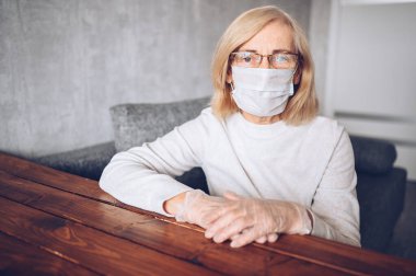 Yalnız, üzgün, yaşlı, koruyucu maskeli, gözlüklü, tıbbi eldivenli yaşlı bir kadın Coronavirus COVID19 salgını sırasında evde karantinada oturuyor. Evde kal konsepti