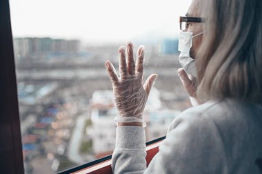 Medikal maskeli ve eldivenli yaşlı yalnız yaşlı kadın kendini karantinaya almak için evde yalnız kalıyor. Ev karantinası, COVID-19 önleme, Coronavirüs salgını durumu
