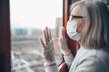 Medikal maskeli ve eldivenli yaşlı yalnız yaşlı kadın kendini karantinaya almak için evde yalnız kalıyor. Ev karantinası, COVID-19 önleme, Coronavirüs salgını durumu