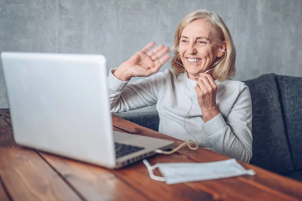 Technology, old age and people concept - happy older senior woman with face medical mask working and making a video call with laptop computer at home during coronavirus COVID19 pandemic. Stay home