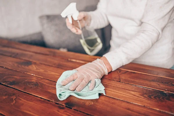 Close up of cleaning home wood table, sanitizing kitchen table surface with disinfectant antibacterial spray bottle, washing surfaces with towel and gloves. COVID-19 prevention sanitizing inside.