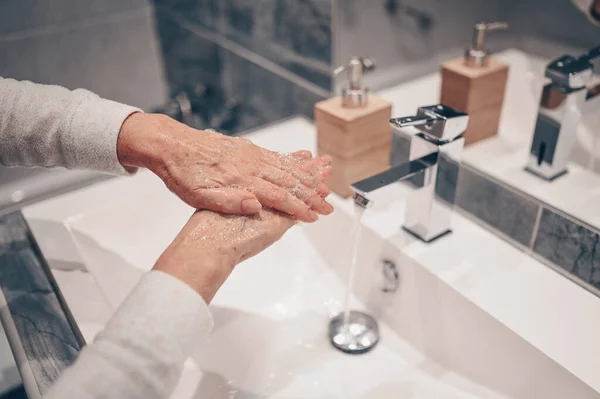 Hand washing lather liquid soap rubbing wrists handwash step senior woman rinsing in water at bathroom faucet sink. Wash hands for COVID-19 spreading prevention. Coronavirus pandemic outbreak.