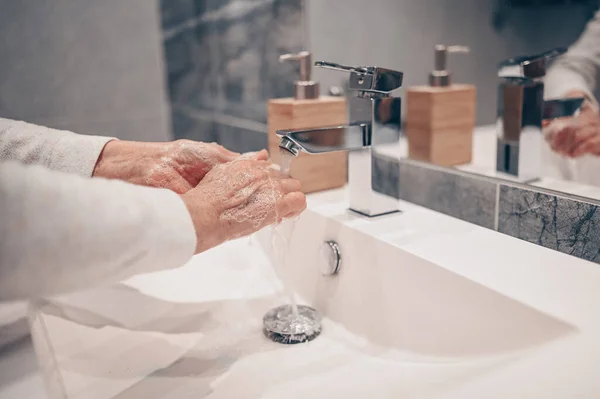 Hand washing lather liquid soap rubbing wrists handwash step senior woman rinsing in water at bathroom faucet sink. Wash hands for COVID-19 spreading prevention. Coronavirus pandemic outbreak.