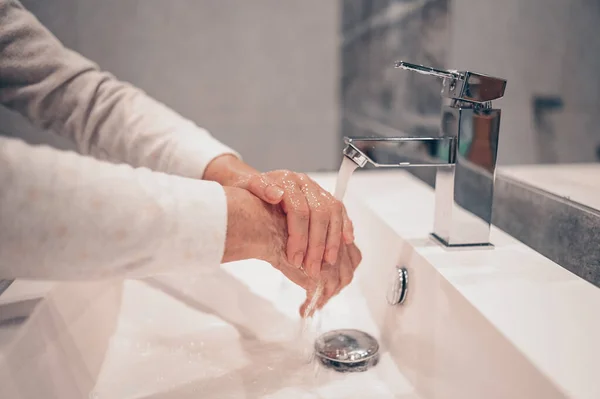 Hand washing lather liquid soap rubbing wrists handwash step senior woman rinsing in water at bathroom faucet sink. Wash hands for COVID-19 spreading prevention. Coronavirus pandemic outbreak.