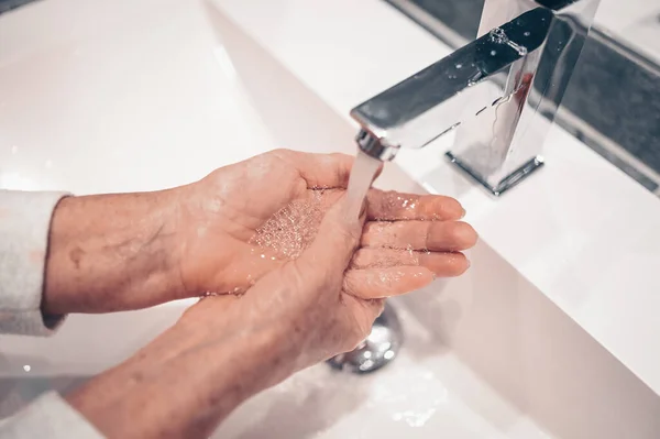 Hand washing lather liquid soap rubbing wrists handwash step senior woman rinsing in water at bathroom faucet sink. Wash hands for COVID-19 spreading prevention. Coronavirus pandemic outbreak.