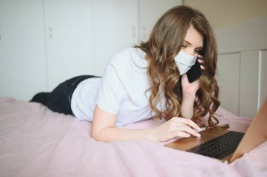 Yüz maskesi takan genç bir kadın telefonda konuşuyor ve Coronavirus karantinası sırasında dizüstü bilgisayarla yatakta çalışıyor. Covid-19 salgını Corona virüsü. Ev kavramından uzaklık çevrimiçi işi.