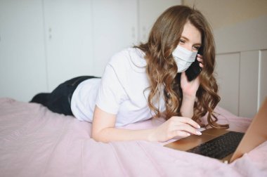 Yüz maskesi takan genç bir kadın telefonda konuşuyor ve Coronavirus karantinası sırasında dizüstü bilgisayarla yatakta çalışıyor. Covid-19 salgını Corona virüsü. Ev kavramından uzaklık çevrimiçi işi.