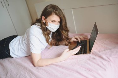 Coronavirus ev karantinası sırasında yüz maskeli genç Avrupalı kadın dizüstü bilgisayar ve yatakta çalışıyor. Covid-19 salgını Corona virüsü. Ev kavramından uzaklık çevrimiçi işi.