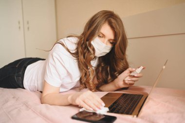 Coronavirus karantinası sırasında hasta kadın el dezenfektanı ile ev temizleme laptopunu karantina altında tutuyor, pamuk yünü ve alkol kullanıyor. Coronavirus COVID-19 salgını. Güvenli ev konsepti