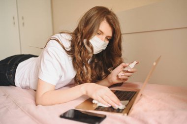 Coronavirus karantinası sırasında hasta kadın el dezenfektanı ile ev temizleme laptopunu karantina altında tutuyor, pamuk yünü ve alkol kullanıyor. Coronavirus COVID-19 salgını. Güvenli ev konsepti