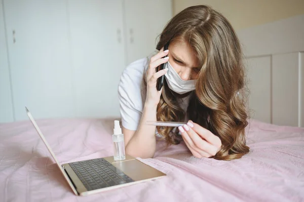 Sick woman in face protection mask lying in bed with laptop and sanitiser holding thermometer at home quarantine isolation. Distance online work from home. Corona virus infection COVID-19 concept.