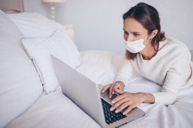 Coronavirus ev karantinası sırasında yatak odasında dizüstü bilgisayarda çalışan yüzü tıbbi maskeli genç Avrupalı bir kadın. Covid-19 salgını Corona virüsü. Ev kavramından uzaklık çevrimiçi işi.