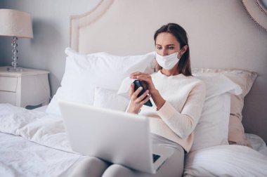 Genç Avrupalı kadın yatak odasında dizüstü bilgisayarla koronavirüs karantinası sırasında el dezenfektanı ile ev temizleme telefonu kullanıyor ve pamuk ve alkol kullanıyor. Coronavirus COVID-19 salgını