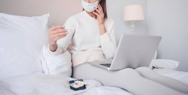 Yüz koruma maskeli tanımlanamayan hasta bir kadın dizüstü bilgisayarında termometre ve haplarla evde karantina hücresinde yatıyor. Evden internet işi. Corona virüs enfeksiyonu COVID-19 konsepti.