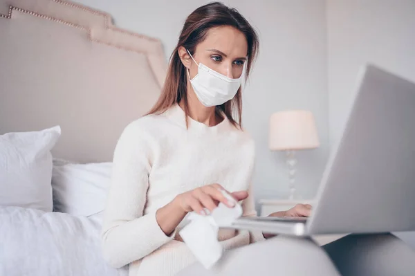European woman in face mask in bedroom during coronavirus isolation home quarantine cleaning laptop by hand sanitizer, using cotton wool with alcohol to wipe to avoid contaminating with Corona virus.