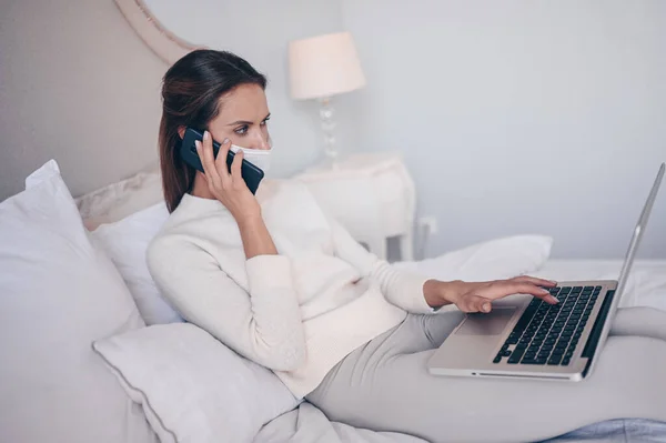 Young woman in bed, speaking phone and working on a laptop. Home quarantine self isolation during pandemic Corona virus. Distance work from home. COVID-19 concept to promote stay safe home save lives