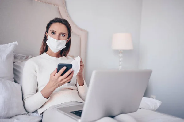Young european woman in face mask in bedroom with laptop during coronavirus isolation home quarantine cleaning phone by hand sanitizer, using cotton wool with alcohol. Coronavirus COVID-19 pandemic