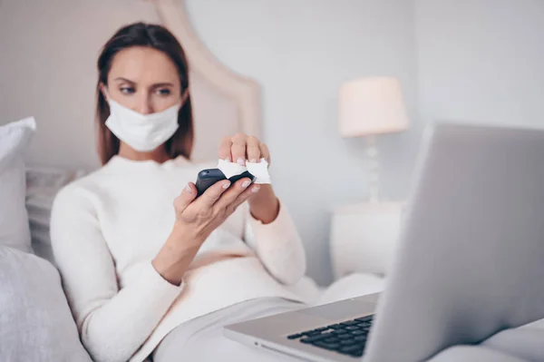 Young european woman in face mask in bedroom with laptop during coronavirus isolation home quarantine cleaning phone by hand sanitizer, using cotton wool with alcohol. Coronavirus COVID-19 pandemic