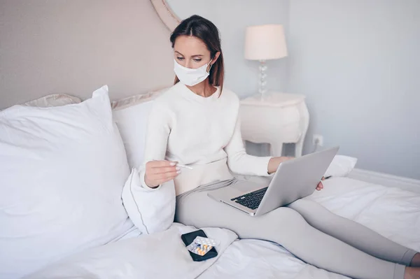 Sick woman in face protection mask lying in bed holding thermometer at home quarantine isolation. Stay home safe concept. Suspecting Corona virus infection COVID-19. First symptoms fever and cough