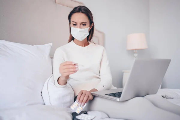 Sick woman in face protection mask lying in bed holding thermometer and pills at home quarantine isolation. Corona virus COVID-19 concept. Suspecting Infection. Respiratory illness first symptom fever