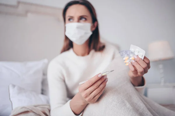 Sick woman in face protection mask lying in bed holding thermometer and pills at home quarantine isolation. Corona virus COVID-19 concept. Suspecting Infection. Respiratory illness first symptom fever