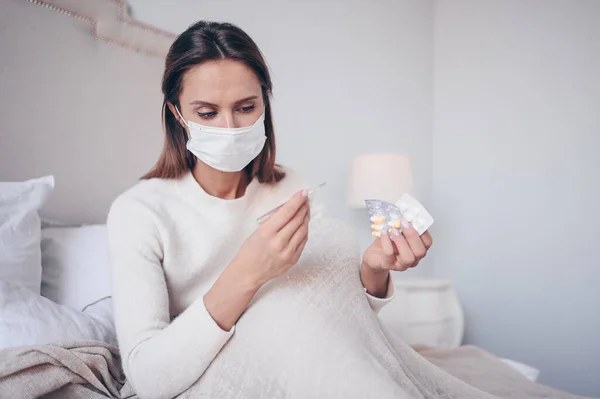 Sick woman in face protection mask lying in bed holding thermometer and pills at home quarantine isolation. Corona virus COVID-19 concept. Suspecting Infection. Respiratory illness first symptom fever