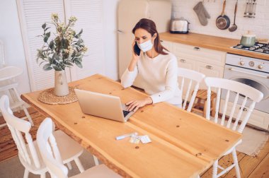 Tıbbi maskeli Avrupalı hasta bir kadın akıllı telefon konuşuyor ve mutfakta dizüstü bilgisayar üzerinde çalışıyor. Covid-19 pandemik Corona virüsü. Çevrimiçi iş, evde kal. Termometre ve haplar
