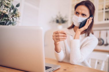 Medikal maskeli Avrupalı hasta bir kadın elinde akıllı telefon ve termometreyle dizüstü bilgisayarla mutfak karantinası Covid-19 pandemik Corona virüsü üzerinde çalışıyor. Mesafe çevrimiçi iş, evde kal