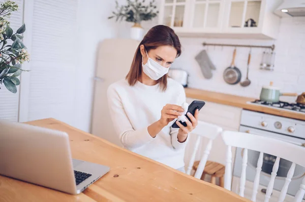 woman in face protection mask cleaning mobile phone to eliminate germs Covid-19 by hand sanitizer, using cotton wool with alcohol to wipe to avoid contaminating with Corona virus.