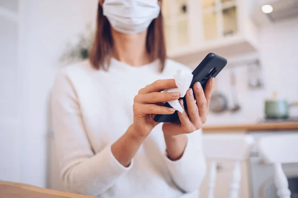 Unrecognizable woman in face protection mask cleaning mobile phone to eliminate germs Covid-19 by hand sanitizer, using cotton wool with alcohol to wipe to avoid contaminating with Corona virus.