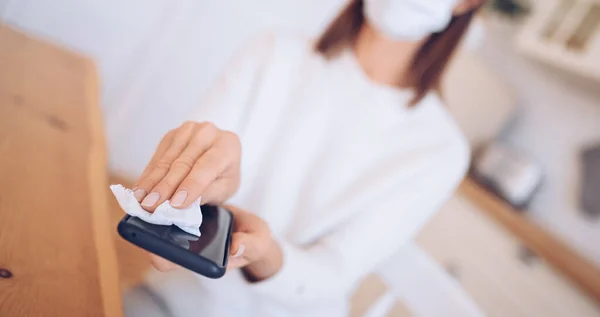 Unrecognizable woman in face protection mask cleaning mobile phone to eliminate germs Covid-19 by hand sanitizer, using cotton wool with alcohol to wipe to avoid contaminating with Corona virus.