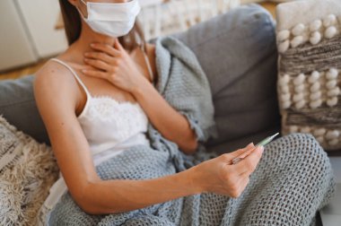 Kanepede yüzü tanınmayan hasta bir kadın, gri, rahat battaniyeli, termometresi olan, kendini karantinaya almış. Corona virüsü enfeksiyonu. COVID-19 konsepti güvenli ev yaşamını teşvik ediyor.