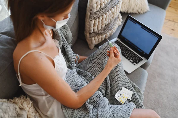 Sick freelance woman in face protection mask sitting on couch in blanket with laptop holding thermometer and pills, home quarantine self isolation. Distance online work from home. Corona virus infection COVID-19 concept. Remote work from home