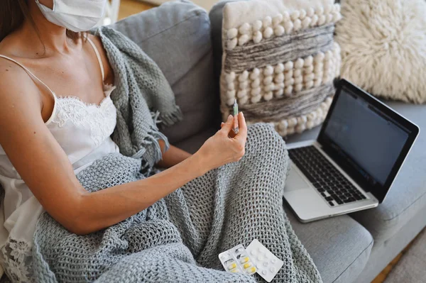 Sick freelance woman in face protection mask sitting on couch in blanket with laptop holding thermometer and pills, home quarantine self isolation. Distance online work from home. Corona virus infection COVID-19 concept. Remote work from home