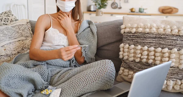 Sick unrecognizable woman in face protection mask on couch with grey comfortable blanket holding thermometer, home quarantine self isolation. Corona virus infection. COVID-19 concept promote stay safe home save lives