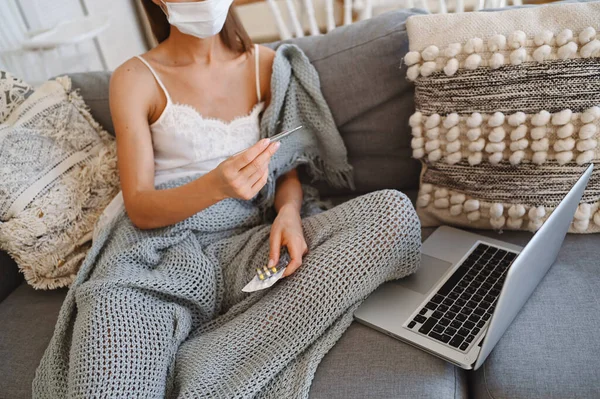 Sick unrecognizable woman in face protection mask on couch with grey comfortable blanket holding thermometer, home quarantine self isolation. Corona virus infection. COVID-19 concept promote stay safe home save lives. Remote work from home
