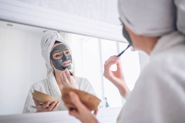 Kozmetik, cilt bakımı, yüz bakımı, spa, doğal güzellik konsepti. Bornozlu güzel bir kadın, sorunlu cildini tazelemek için yüz kilinden maske takıyor.